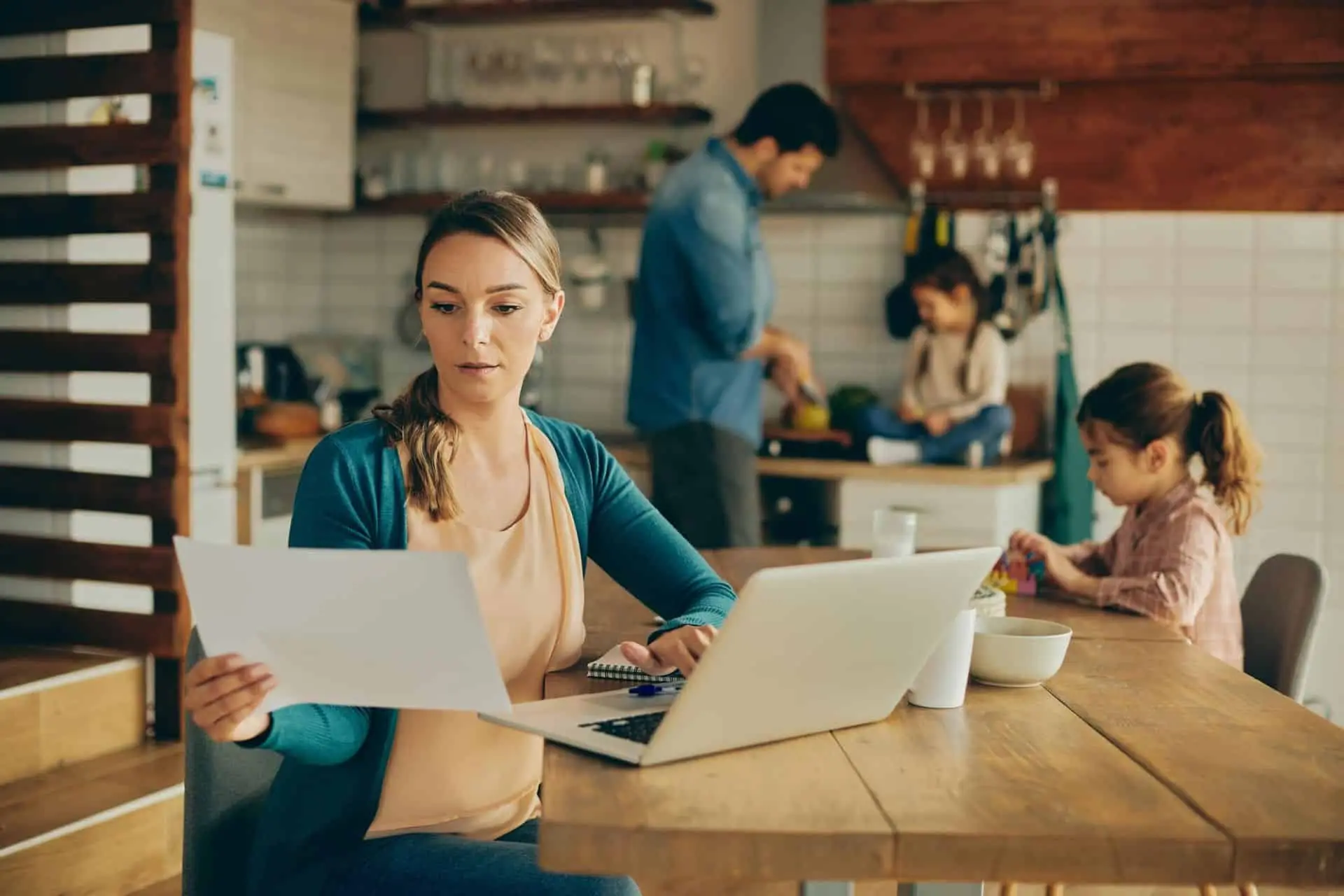 A woman reviewing papers at a kitchen table while her family is in the kitchen