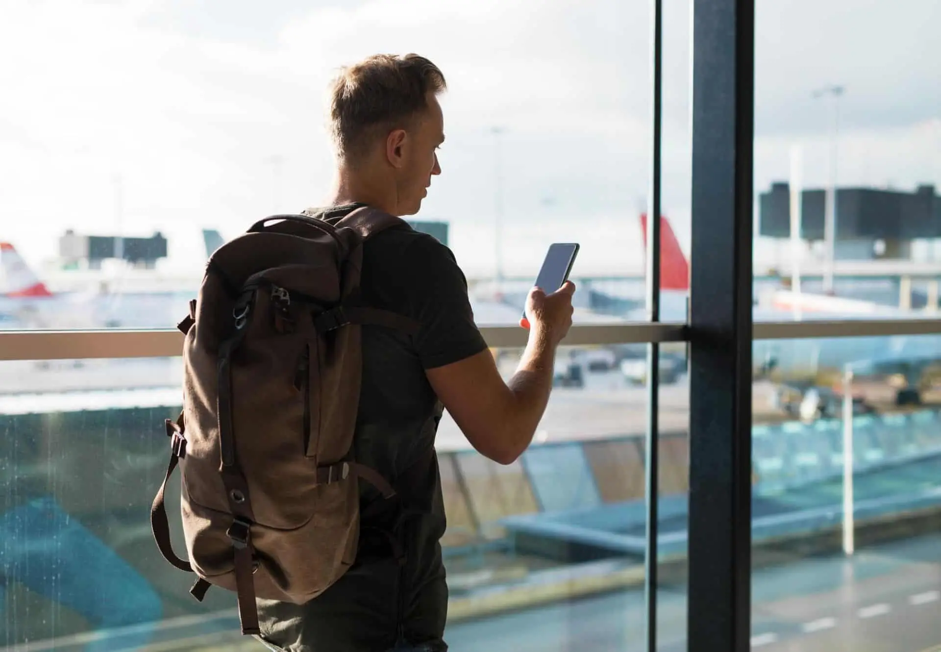A man with a backpack using his phone at an airport window