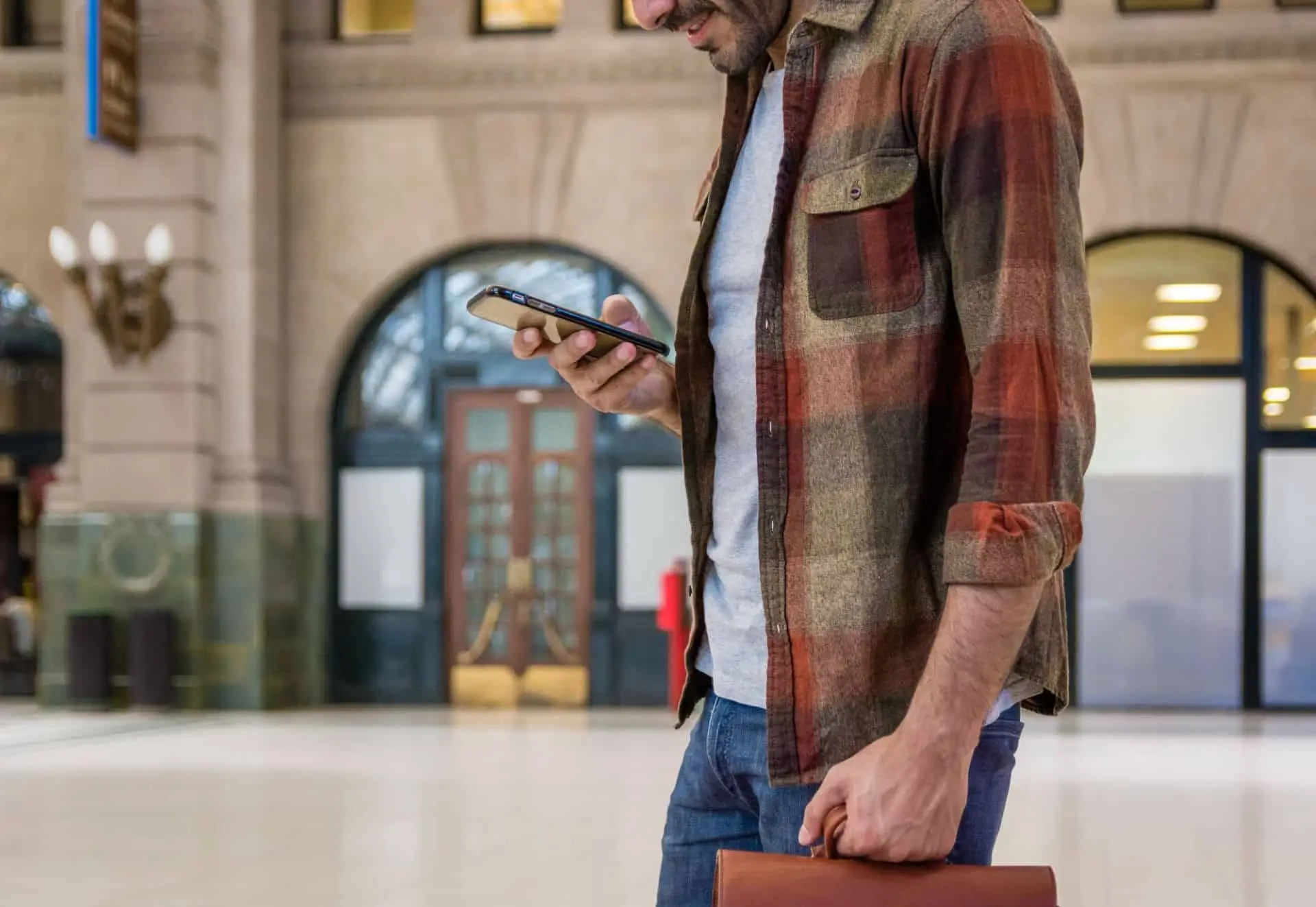 A man checking his phone in a station hall with a bag in hand